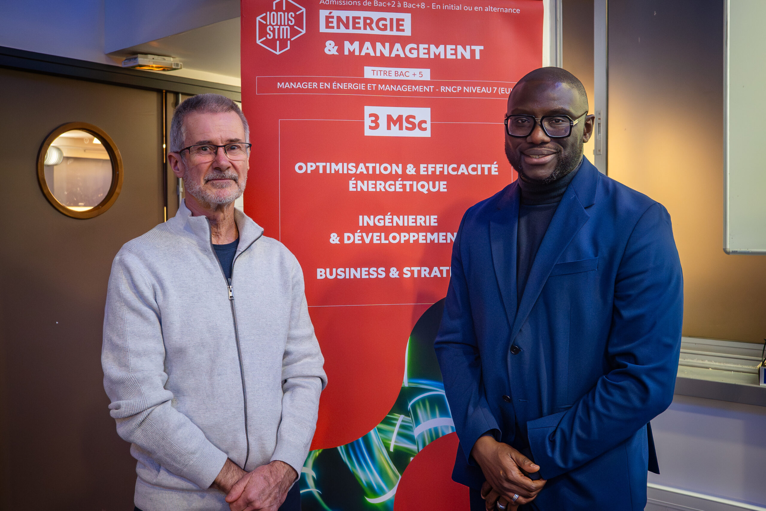 Deux personnes sourient devant un kakémono rouge, ce sont des hommes